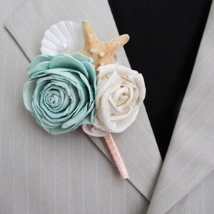 May include: A boutonniere featuring a light teal rose, a cream-colored rose, a starfish, and a seashell. The floral arrangement is accented with a pink ribbon and is pinned to a light gray pinstripe suit jacket.