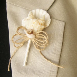 Puede incluir: Un boutonniere de concha con una concha blanca, una concha marrón más pequeña y un lazo de cuerda marrón.
