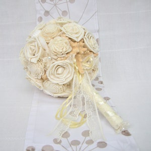 May include: A white and cream bridal bouquet made of seashells and faux flowers with a yellow ribbon and lace trim.