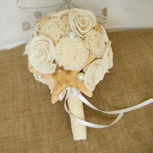 May include: A white and cream bridal bouquet with a starfish, seashells, and pearls. The bouquet is tied with a white satin ribbon.