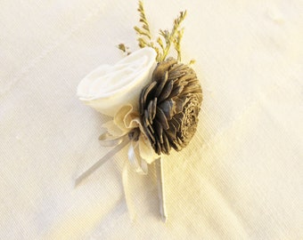 May include: A white and gray floral arrangement with a white rose and a gray pinecone. The arrangement is tied with a white ribbon and has a few sprigs of greenery.
