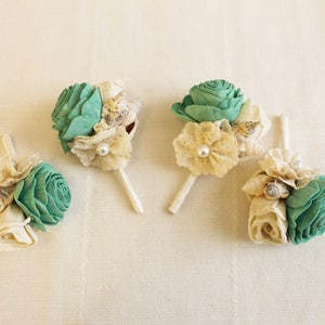 May include: Four small bouquets of teal and white fabric flowers with seashells and pearls. The bouquets are attached to wooden skewers.