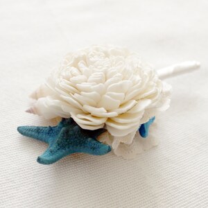 May include: A handcrafted boutonniere featuring a cream-colored flower, a small seashell, and a turquoise starfish. The boutonniere is accented with delicate lace and a white satin stem, perfect for a beach-themed wedding or event.