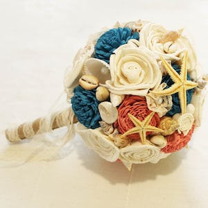May include: A white, coral, and teal bridal bouquet with seashells and starfish. The bouquet has a burlap and lace handle.