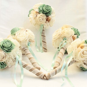 May include: Five bouquets of artificial flowers with a beach theme. The bouquets are made with white and cream roses, green succulents, and seashells. The bouquets are tied with a light green ribbon and have a burlap stem.