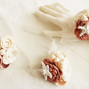 May include: Three decorative wrist corsages with a beach theme. Each corsage features a cluster of white fabric roses, coral-colored flowers, seashells, and a white starfish. The corsages are tied with sheer ribbons.