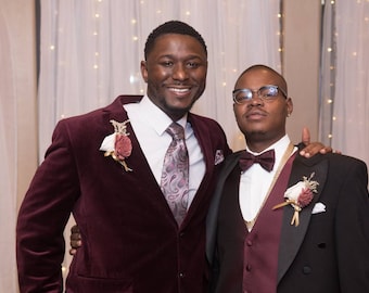 May include: Two men in formal attire. One wears a burgundy velvet blazer with a floral boutonniere, a white shirt, and a patterned tie. The other man wears a black tuxedo with a burgundy vest, bow tie, and a floral boutonniere.
