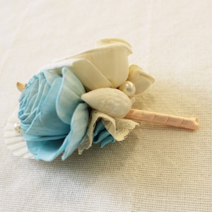 May include: A light blue and white floral boutonniere with a pearl accent and a pink ribbon.