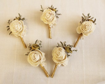 May include: Five ivory-colored paper flower boutonnieres with gold stems and green foliage accents.
