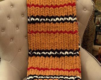May include: A chunky knit blanket in shades of orange, with horizontal stripes of black, white, and red. The blanket is folded over the arm of a beige upholstered armchair, showcasing its texture and colour pattern.