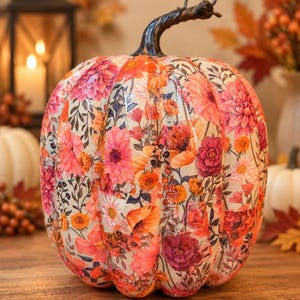 May include: A decorative pumpkin featuring a decoupage floral pattern with vibrant shades of pink, orange, and deep red flowers against a cream background. The pumpkin has a dark, textured, curved stem and a rounded, ribbed shape.
