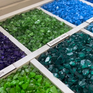 May include: A close-up of six different colours of crushed glass, including green, blue, purple, and teal. The glass is labelled with the name "Bushstone Green".