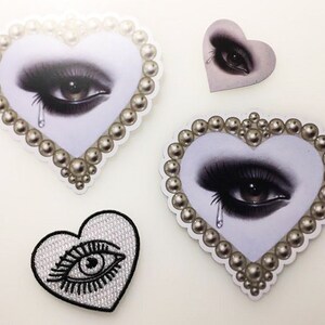 May include: Four heart-shaped patches with a pearl border. Each patch features a black and white illustration of a single eye with a tear. Two of the patches are larger and two are smaller.