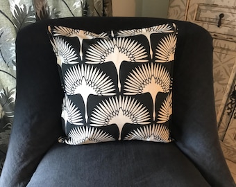 May include: A square decorative pillow with a black background and a repeating white bird pattern. The pillow is placed on a dark gray upholstered chair. The bird design features a stylized fan shape.