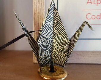 May include: A black and gold origami crane with a decorative pattern, attached to a gold-colored base. A dark ribbon extends from the crane. The crane is on a wooden surface, near a patterned vase and a framed print.