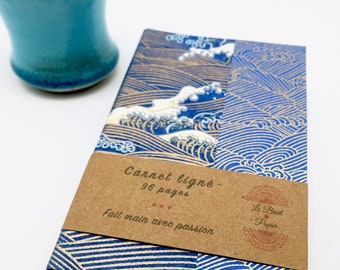 May include: A blue and gold patterned notebook with a matching blue ceramic cup. The notebook has a brown paper band with the text "Carnet ligné - 96 pages" and "Fait main avec passion".