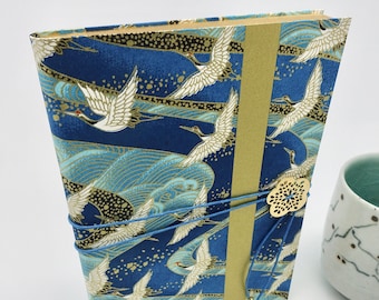 May include: A handcrafted journal with a Japanese-inspired design. The cover features a blue fabric with white cranes in flight over stylized waves. A gold-colored spine and a blue cord closure with a decorative charm complete the design.