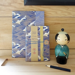 Può includere: Due quaderni con disegni di onde blu e dorate. Il quaderno più piccolo ha una copertina di carta marrone con il testo "Carnet secret 144 pages Papier uni Vrai ivoire Fait main avec passion A Petit Paper" su di essa. Una bambola di legno con un kimono blu e verde è sullo sfondo.
