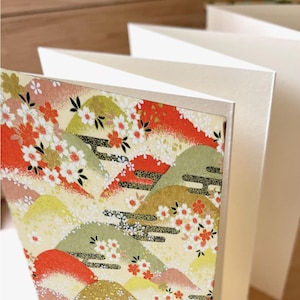 Może przedstawiać: Zbliżenie na zestaw pustych kartek okolicznościowych. Przednia karta ma kolorowy japoński wzór z górami, kwiatami wiśni i kremowym tłem. Pozostałe karty są białe.