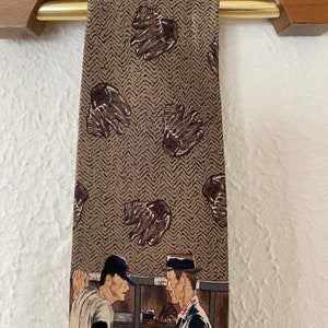 May include: A brown silk tie with a pattern of baseball gloves and a scene of two baseball players in a dugout. The tie has a herringbone pattern.