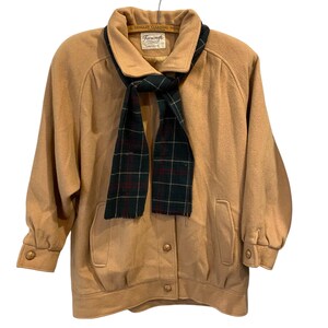 May include: A tan jacket with a matching scarf. The jacket has a collared neckline, two front pockets, and gold buttons. The scarf is green with a red and black plaid pattern. The jacket is hanging on a metal hanger.