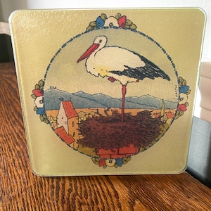 May include: A square glass cutting board featuring a central circular illustration of a white stork with a red beak standing in a nest on a chimney. The scene includes a village with red-roofed buildings, mountains, and a floral border.