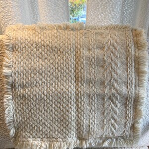 May include: Cream-colored rectangular throw pillow with textured woven patterns and fringe trim. The pillow features three distinct woven designs: a diamond pattern, a cable knit, and a vertical stripe. The fringe trim adds a soft, decorative edge.