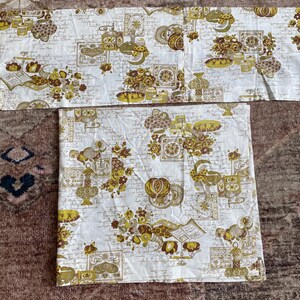 May include: A white fabric with a repeating pattern of brown and gold floral designs. The fabric has a vintage look and feel.