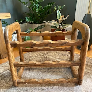 May include: A wooden wine rack featuring a natural grain finish, constructed with two sturdy side panels and three horizontal bars. Each bar contains four scalloped cutouts designed to hold bottles securely in a horizontal position.