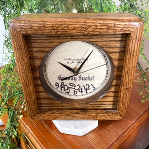 May include: A wooden clock with a white face and black hands. The clock face reads "A Handcrafted Clock by Jim Math" and "Gravity Sucks! 987654321".