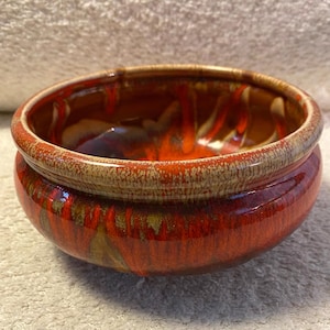 May include: A round ceramic bowl with a glossy, reddish-orange exterior and a lighter, textured rim. The interior features a blend of red, orange, and brown hues. The bowl's design suggests it could be used for serving or as a decorative piece.