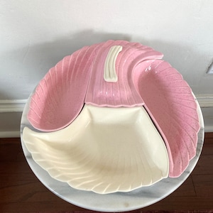 May include: A three-piece serving dish set in a pink and white color scheme. The set features a large, central, white dish with two smaller pink dishes on either side. The dishes have a scalloped edge and a textured surface.
