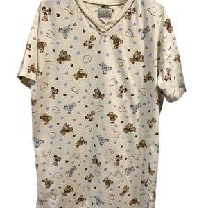 May include: A cream-colored V-neck t-shirt featuring an all-over print of brown and blue teddy bears, outlined hearts, and solid light blue hearts. The inner neck label reads Basic Editions. The shirt has short sleeves and a straight hem.
