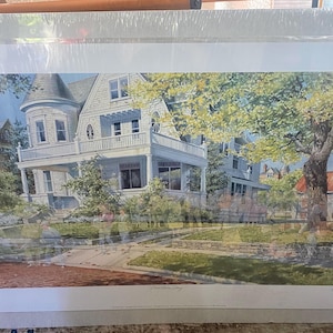 May include: A framed print of a detailed watercolor painting of a neighborhood street scene. The artwork features a large white Victorian house with a wraparound porch, a tree, and other houses. The scene is rendered in soft colors.