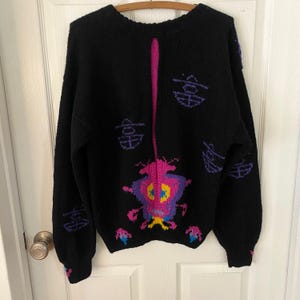 May include: Black knit sweater with a vibrant, colorful design. The sweater features a central graphic of a creature with pink, purple, yellow, and blue accents. Additional purple geometric shapes are scattered across the sweater. A vertical pink stripe runs down the back.