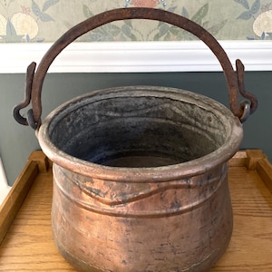 May include: A round copper pot with a weathered, oxidized surface and a wide, open top. The vessel features a thick, dark, rusted iron swing handle attached to two side loops, showing signs of age and heavy use with visible patina.