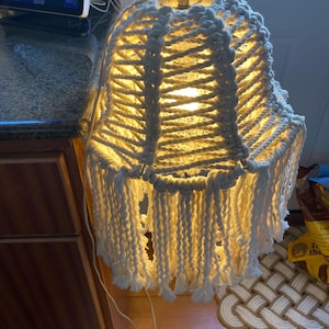May include: A white macrame lampshade with a fringe detail. The lampshade is lit and casts a warm glow.
