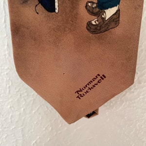 May include: A brown silk tie with a printed image of two children wearing shoes. The tie has the text "Norman Rockwell" embroidered in maroon.