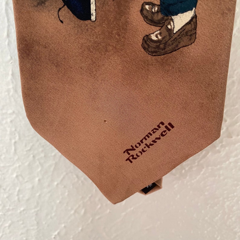 May include: A brown silk tie with a printed image of two children wearing shoes. The tie has the text "Norman Rockwell" embroidered in maroon.