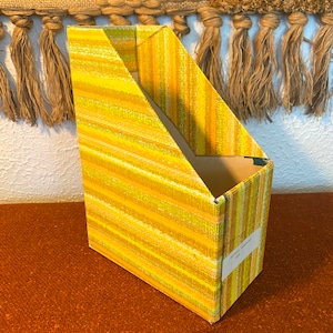 May include: A yellow and orange striped cardboard magazine holder with a single divider. The holder has a label on the side that reads "Wallace & Gromit"
