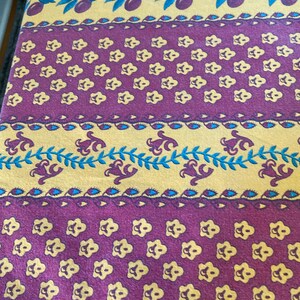 May include: A fabric textile featuring horizontal bands of repeating patterns. The design includes rows of small yellow flowers on a purple background, alternating with yellow stripes containing blue olive branches and stylized purple floral motifs.