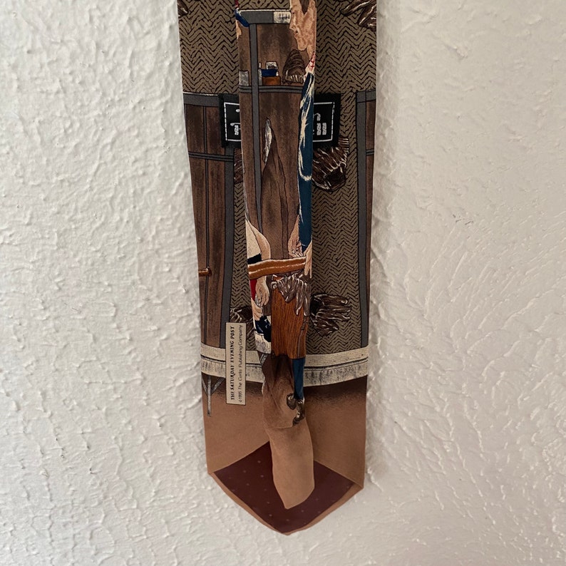 May include: A brown and tan silk tie with a woven design featuring a man in a blue and white uniform holding a tool. The tie has the text "The Little Leather Tie Post" and "The Little Leather Working Company" printed on it.
