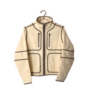 May include: A cream-colored jacket with brown trim and zipper closure. The jacket has a stand-up collar and multiple pockets.