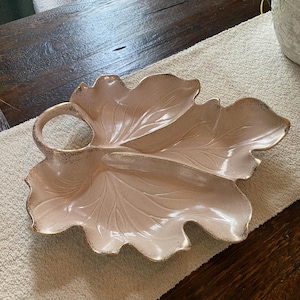 May include: A pink ceramic serving dish shaped like a leaf with three sections and a handle. The dish has a speckled glaze and gold accents.