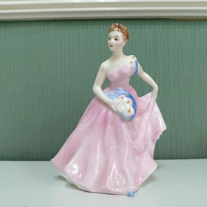May include: A porcelain figurine of a woman in a pink gown, holding a fan. The dress has a flowing design, and the fan is blue with white floral accents. The figurine has a delicate, hand-painted appearance.