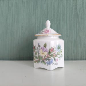 以下が含まれることがあります： 紫、ピンク、青の花柄が施された、蓋付きの白い陶器の瓶。瓶には金の縁取りと蓋に装飾的なつまみが付いています。白い表面に置かれています。