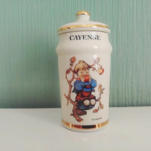 May include: A white ceramic spice jar with a lid, featuring gold trim and the word "CAYENNE" in blue. The jar is decorated with a colorful illustration of a child eating an apple. A charming kitchenware item.