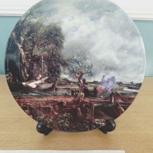 May include: A decorative plate featuring a landscape painting with a boat on the water, trees, and a cloudy sky. The plate is displayed on a black stand, set against a light brown wooden surface.