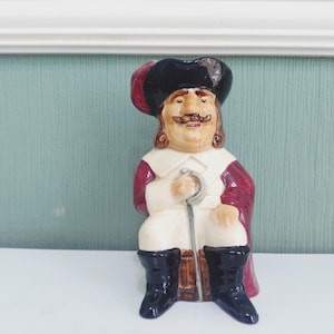 Op de afbeelding: Een keramische karakterkan met een zittende figuur die een zwarte driekante hoed, een witte kraag en een donkerrode jas draagt. De figuur heeft een bruine snor en houdt een zilverkleurige wandelstok vast. Het personage draagt zwarte laarzen en zit op een vat.