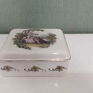 Pode incluir: Uma caixa de porcelana branca com acabamento dourado e um design floral com um casal em uma cena de jardim.